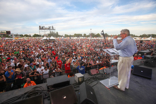 Large crowd of many people standing and watching a person give a speech on a stage