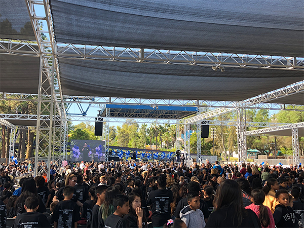Crowds of people standing under sun coverings with large screens in the back